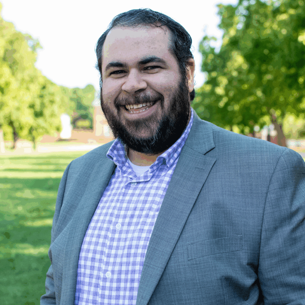 A person in a grey suit and checkered shirt smiles while standing outdoors on a sunny day with green trees in the background.