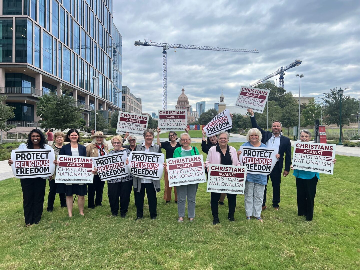 People are standing on grass holding signs with buildings and a crane in the background.