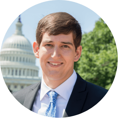 A smiling person in a suit and tie stands outdoors with the US Capitol building in the background.