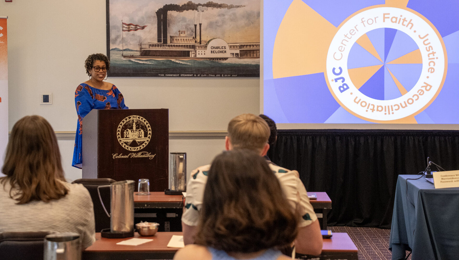 A person speaks at a podium in a conference room with a presentation slide displayed on a screen.