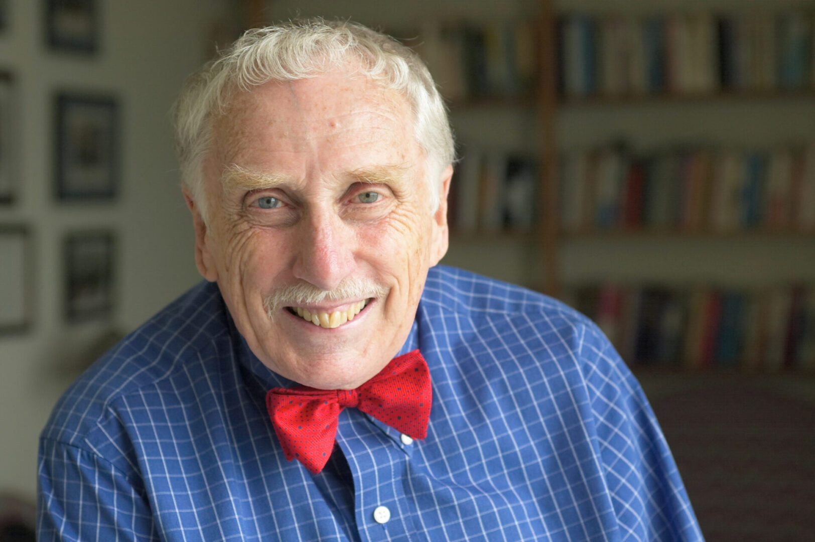 An elderly man with a cheerful expression wears a red bow tie and a blue checkered shirt, standing in a room with a blurred bookshelf in the background.