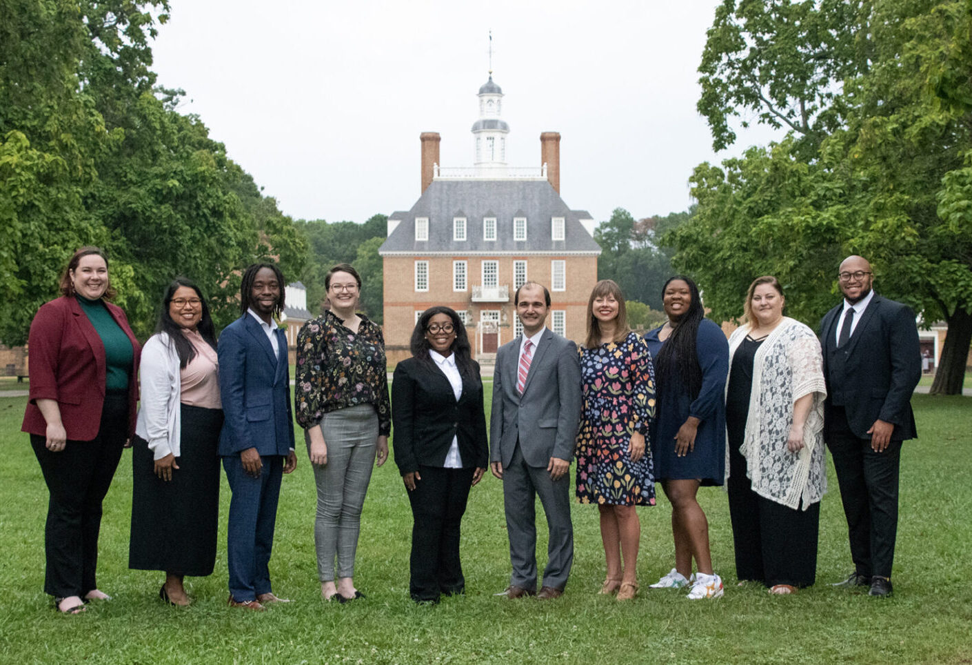 A group of people in formal attire stands outdoors on a lawn with a historic building in the background.