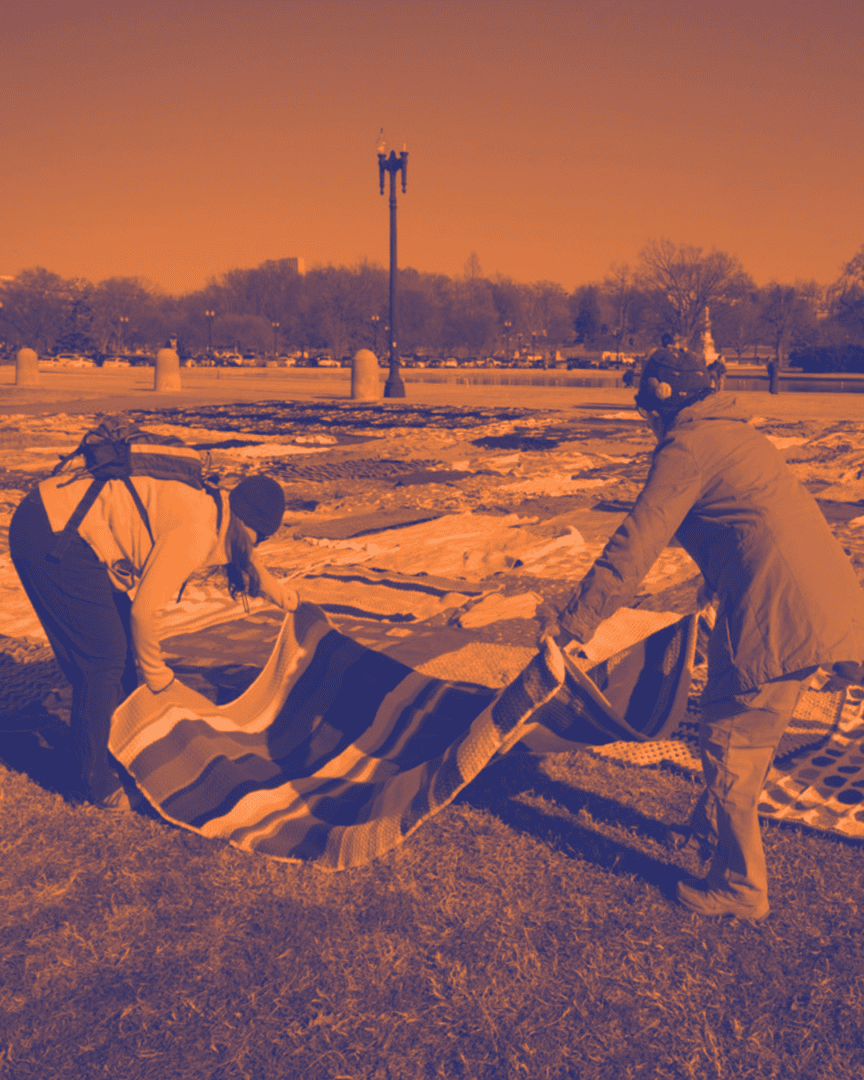 Two people pick a large blanket off the ground, with many other blankets covering the ground behind them.