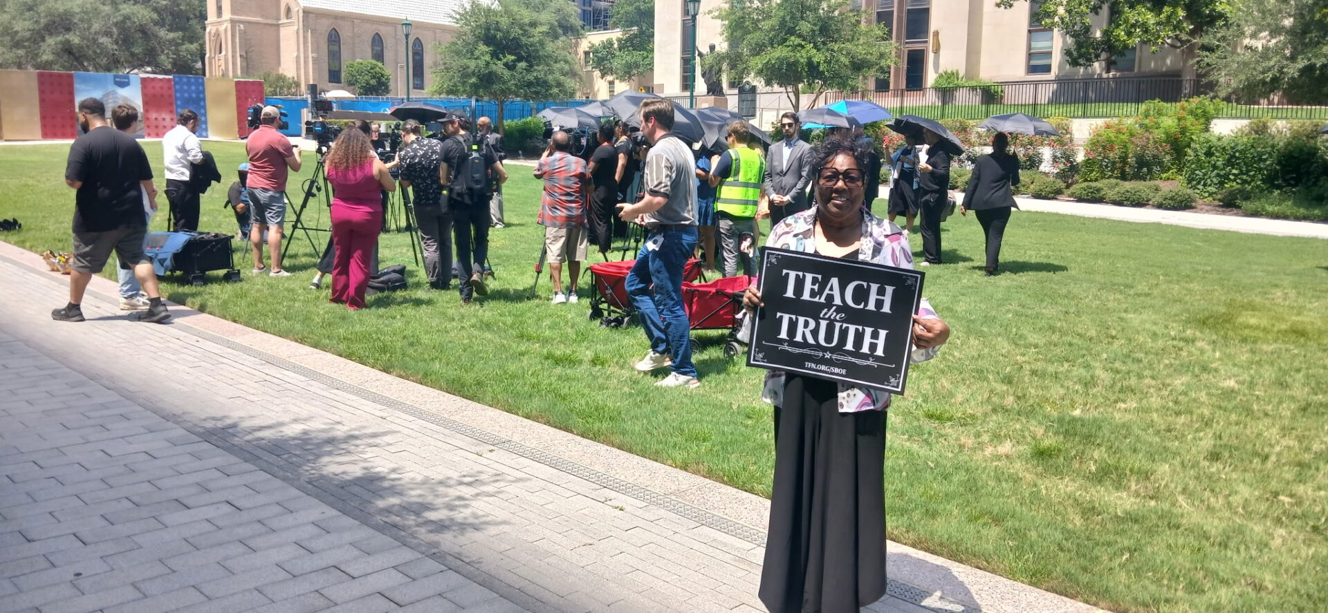 A person holds a "Teach Truth" sign in a gathering outside on a sunny day.